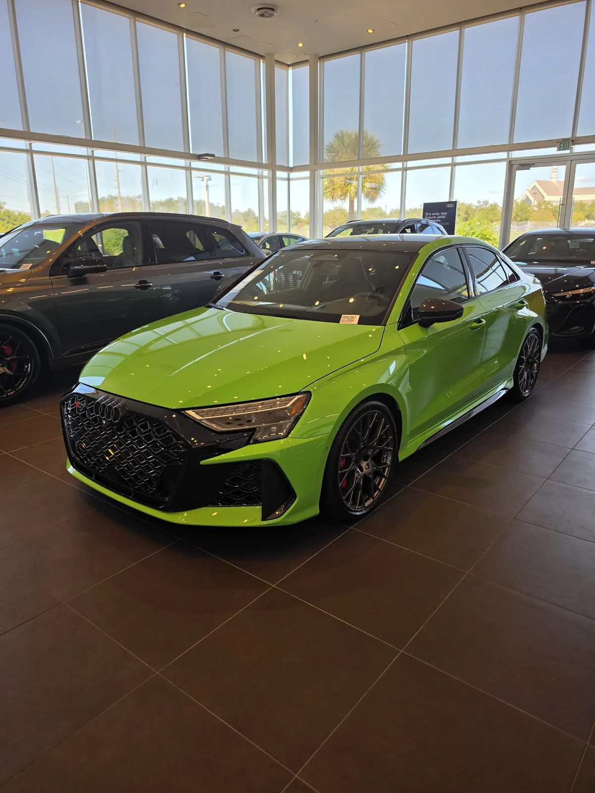 Lime green performance sedan with dark wheels and red brake calipers on a dealership showroom floor, with floor-to-ceiling windows behind it